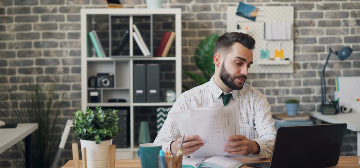 Man reviewing printed papers at a desk with a laptop